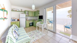 A compact kitchen with a dining area, a sofa, and a view of the sea through a sliding glass door.