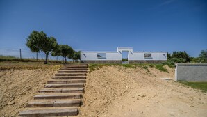 Un edificio moderno con una escalera que lleva a una puerta, rodeado por un paisaje arenoso y vegetación escasa.