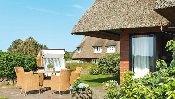 A thatched-roof house with a brick exterior, a patio with wicker furniture, and a garden with various plants.