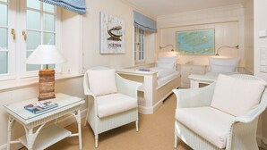 Een kamer met een bed, twee witte rieten stoelen, een kleine tafel met een lamp, een kaart aan de muur, en een raam met een geruit gordijn.