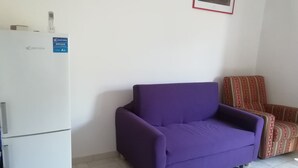 Een paarse bank, een gestreept fauteuil en een witte koelkast in een kamer met witte wanden en een betegelde vloer.