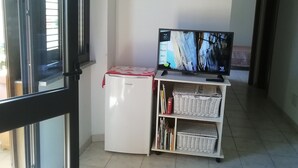 Een kleine witte koelkast met een rode doek erboven, een televisie op een standaard en een rieten mandje op een plank.
