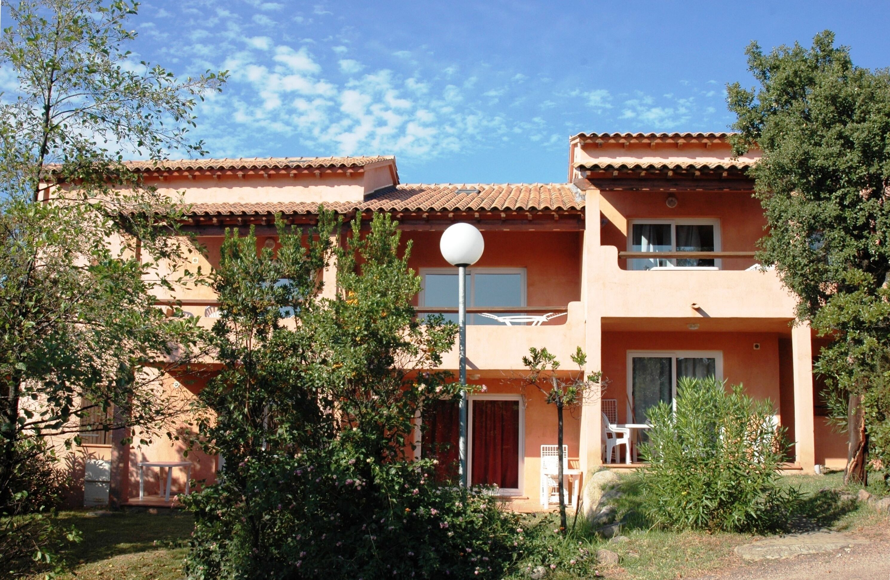 A two-story building with a tiled roof, a balcony, and a small garden area with a white chair and table.