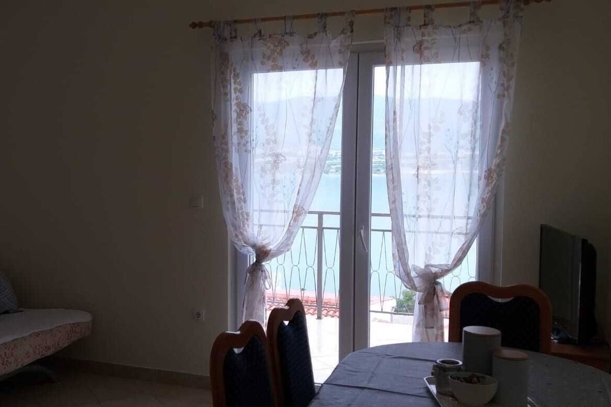 Una habitación con sofá, una mesa de comedor con sillas, televisor y vistas a través de una ventana con cortinas.