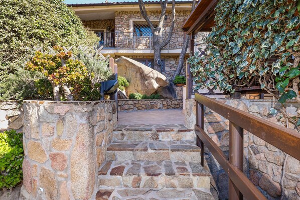 A stone staircase leading to a stone building with a balcony, surrounded by greenery and a wooden railing.