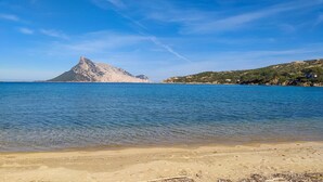 Een strand met helderblauw water, een berg op de achtergrond en een glooiend landschap.