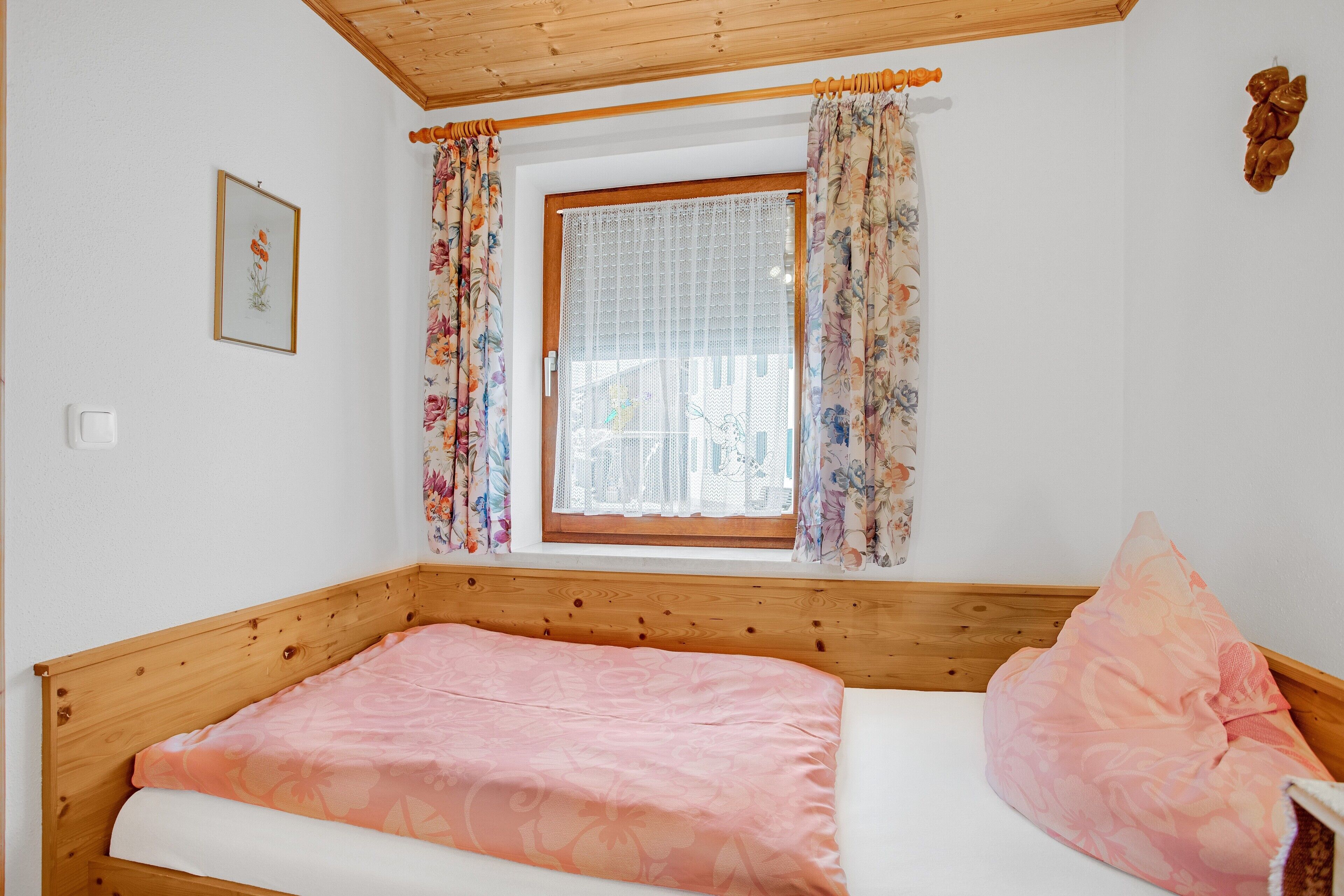A single bed with a wooden headboard, a window with floral curtains, and a framed picture on the wall.