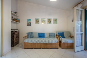 A room with a wicker sofa, a wicker chair, a small shelf with decorative items, and a wall with framed pictures.