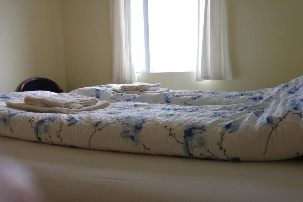 Una cama individual con edredón y almohadas estampadas con flores, una mesita de noche con lámpara, y una ventana con cortinas.