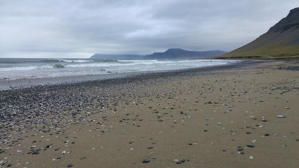 Une plage de galets et de sable, un rivage rocheux et, au loin, des montagnes sous un ciel nuageux.