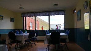 A dining area with tables and chairs, large windows with a view of a red building and a truck, and a clock on the wall.