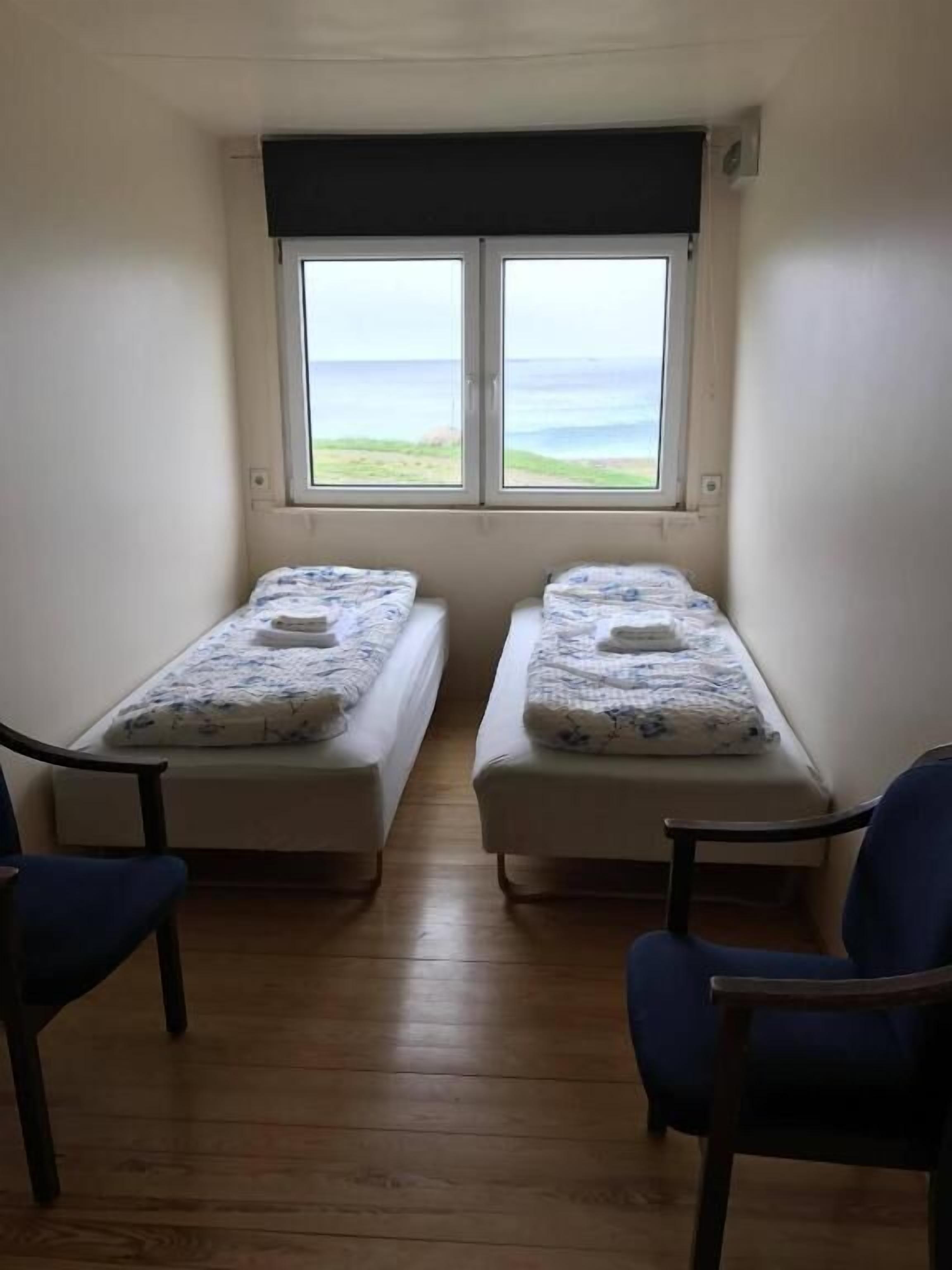 A small room with two single beds, a window with a view of the sea, and two blue chairs.