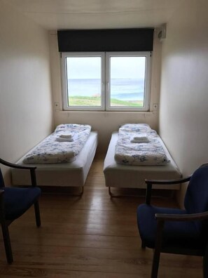 A small room with two single beds, a window with a view of the sea, and two blue chairs.