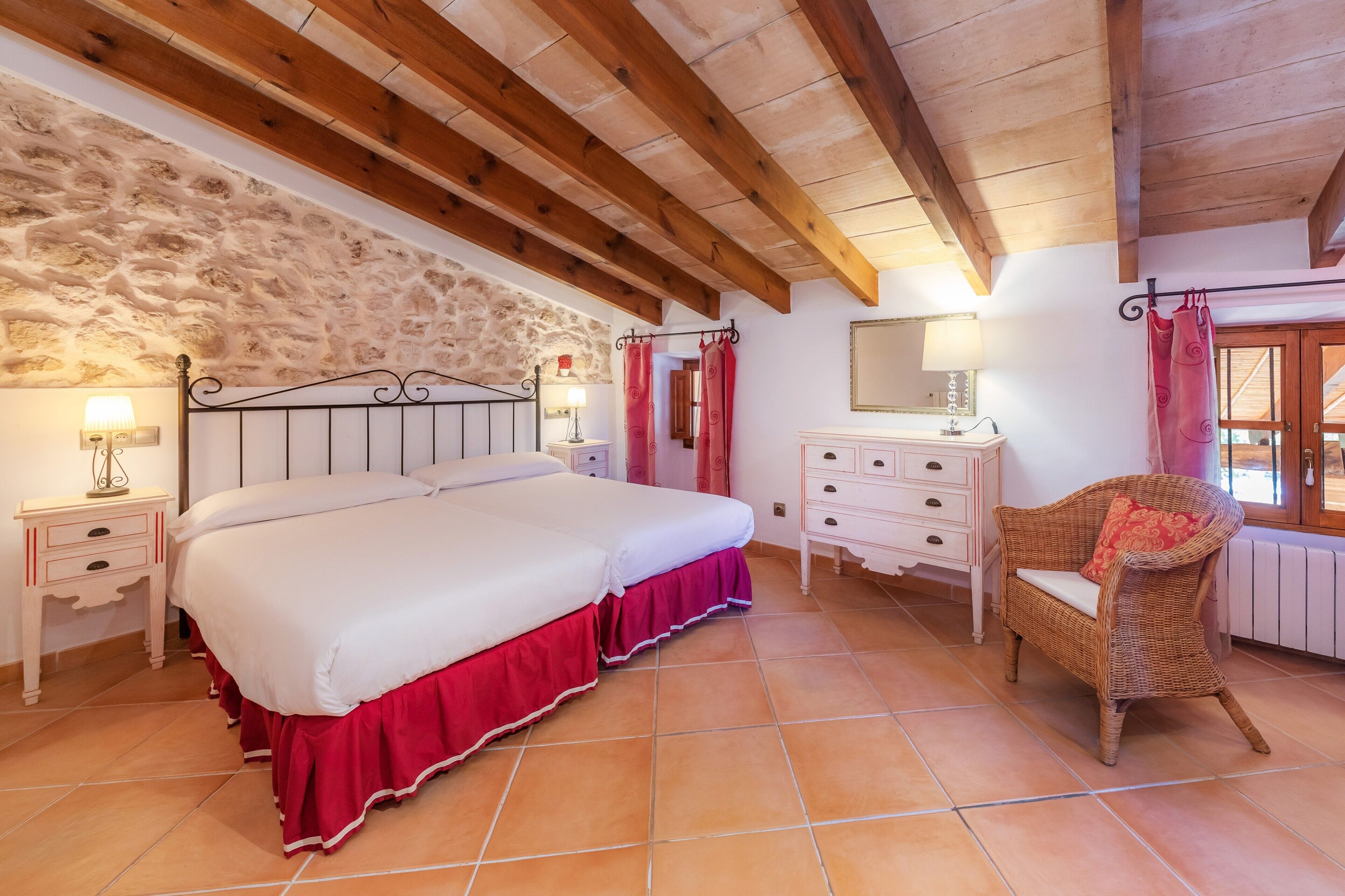 Un dormitorio con una cama grande, vigas de madera, pared de piedra, suelo de baldosas, cómoda, silla y una ventana con cortinas.