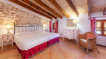 Un dormitorio con una cama grande, vigas de madera, pared de piedra, suelo de baldosas, cómoda, silla y una ventana con cortinas.