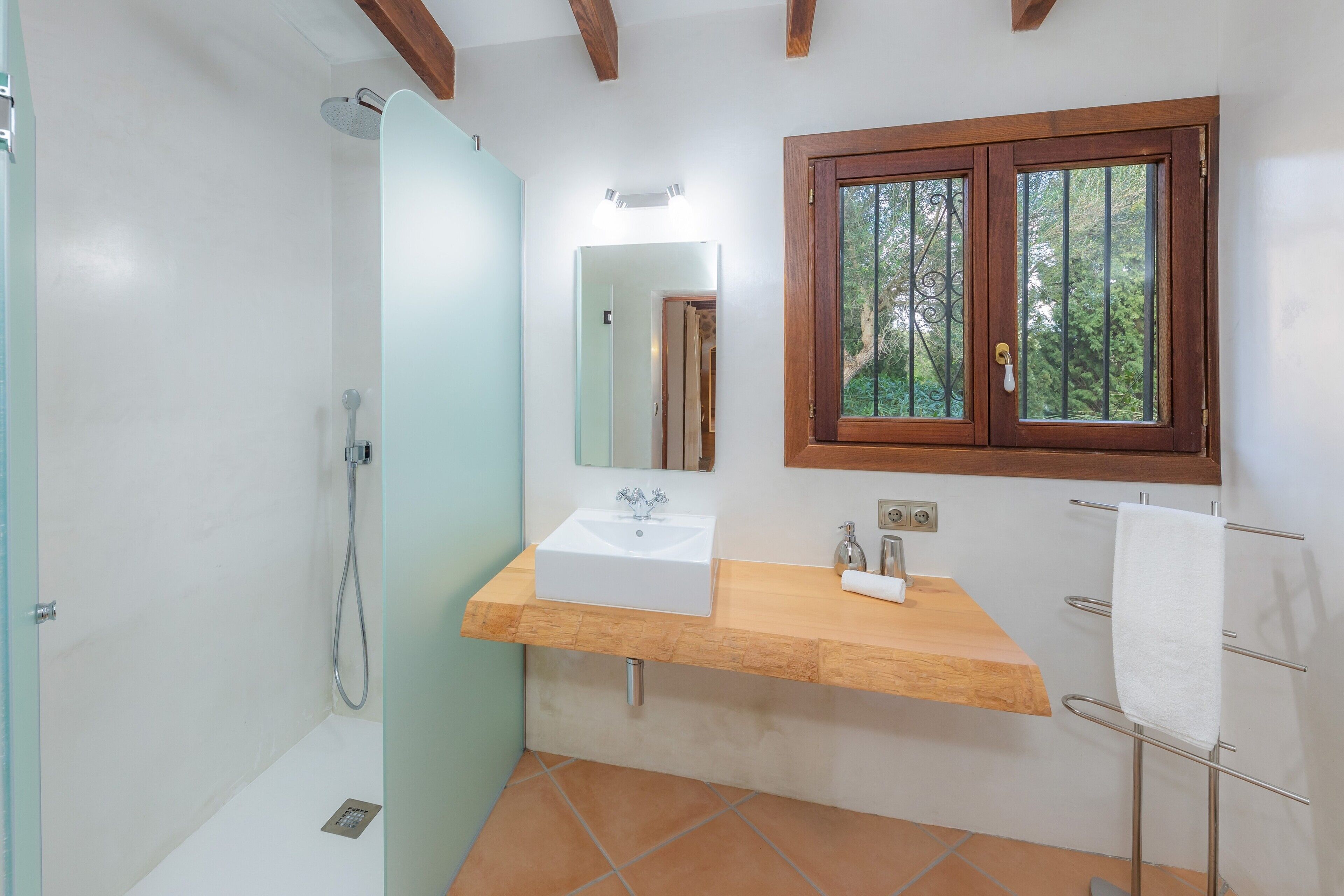 A bathroom with a wooden vanity, a white sink, a glass shower enclosure, and a window with a view of trees.