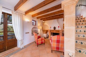 A room with wooden beams, a fireplace, two plaid chairs, a tiled floor, and a view through a glass door.