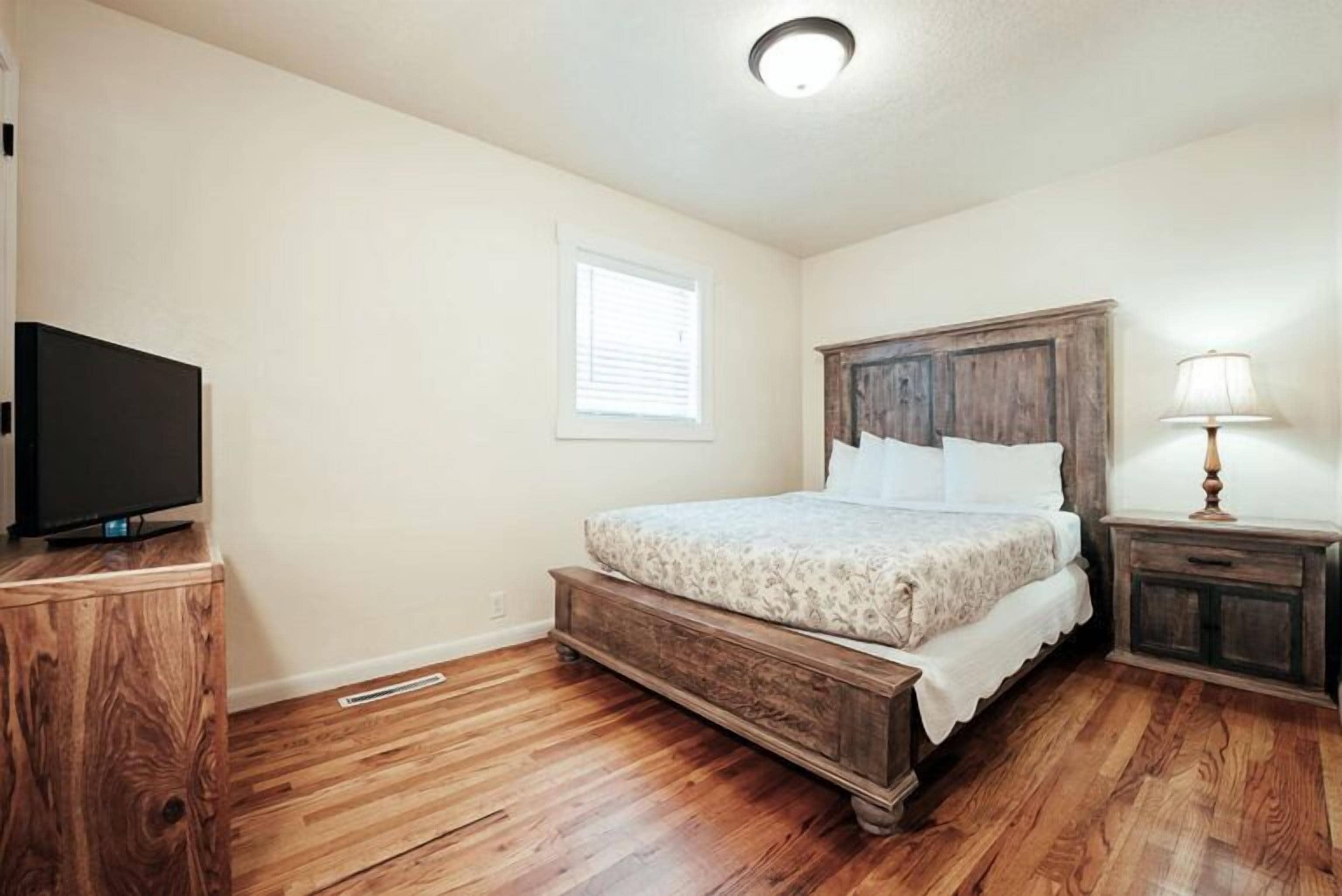 A bedroom with a wooden bed, a television on a wooden cabinet, and a window with blinds.