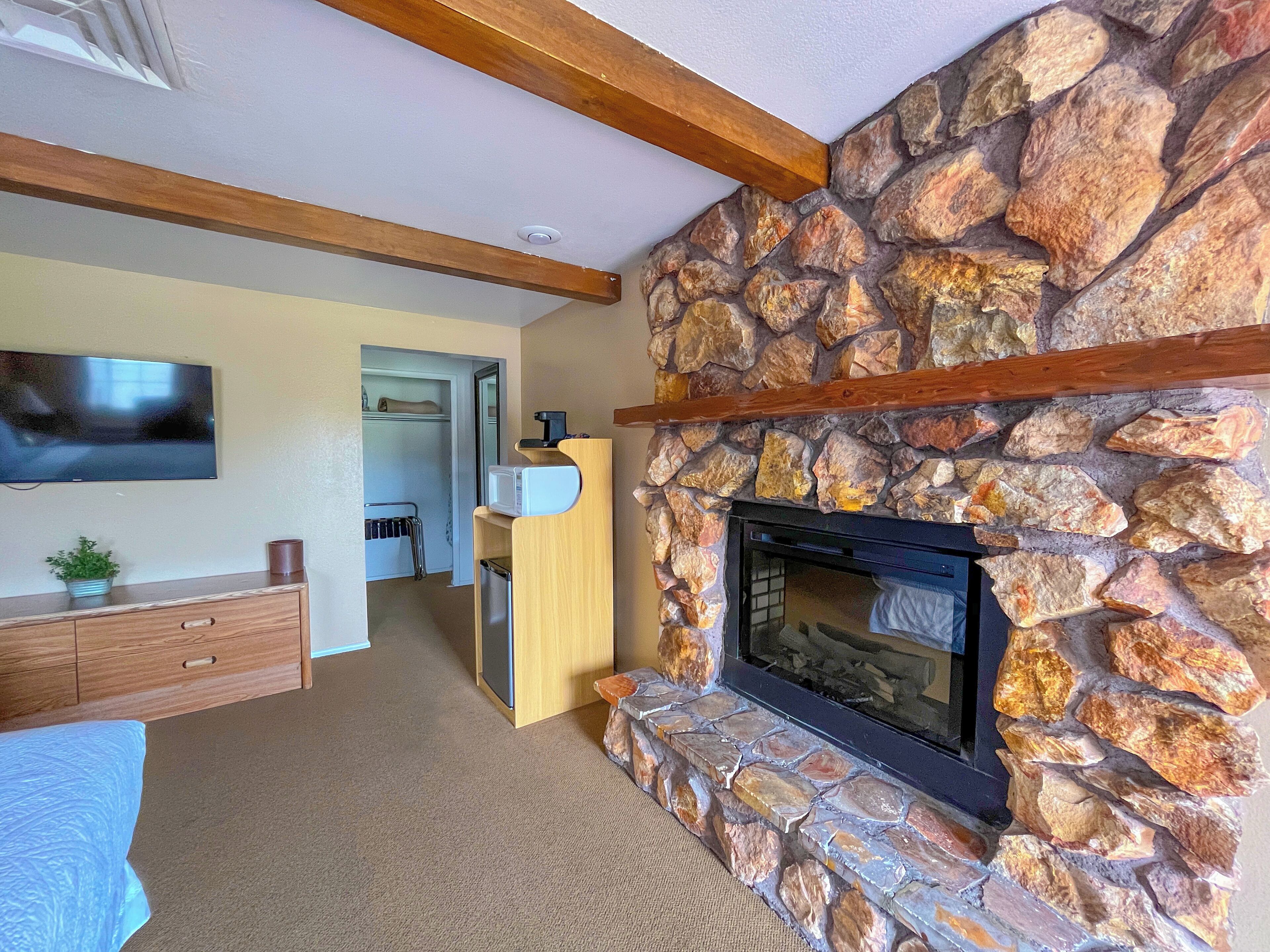 A living room with a stone fireplace, wooden beams, a flat-screen TV, and a kitchenette area.