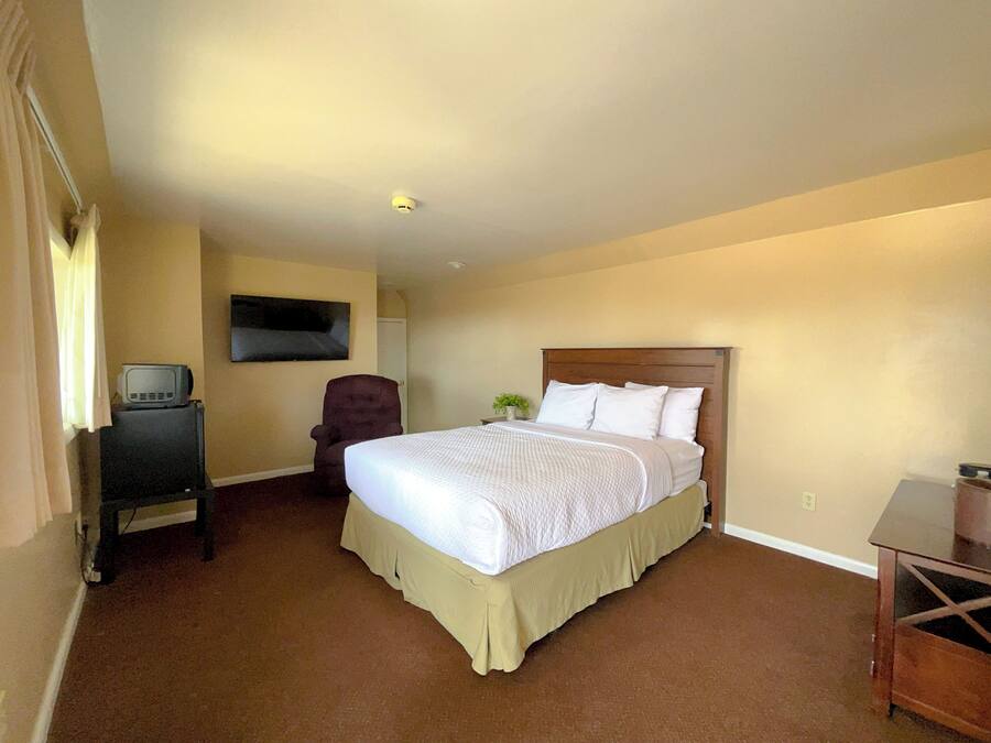 Quarto de hotel com cama grande, uma cadeira, televisão em suporte e uma mesinha.
