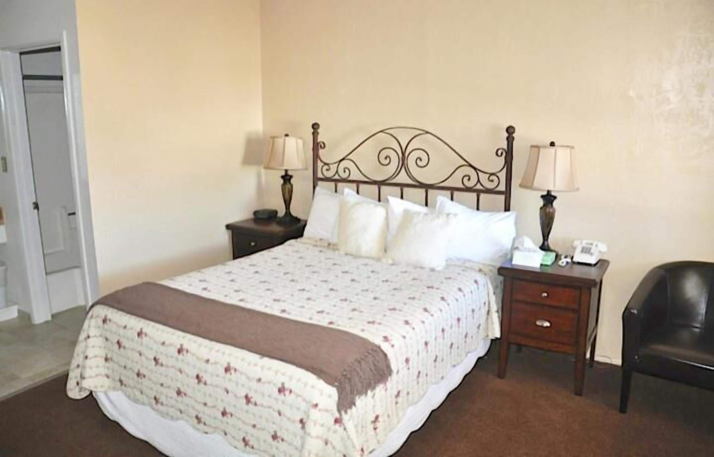 A neatly made bed with a patterned bedspread, two bedside tables with lamps, a chair, and a door in the background.