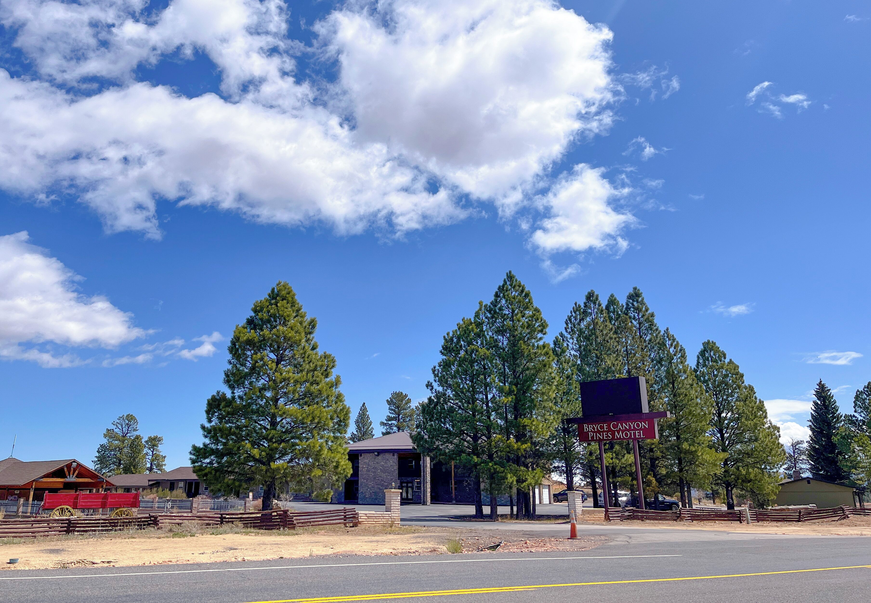 Um motel com uma placa indicando "Bryce Canyon Pines Motel", rodeado por árvores e um céu claro.