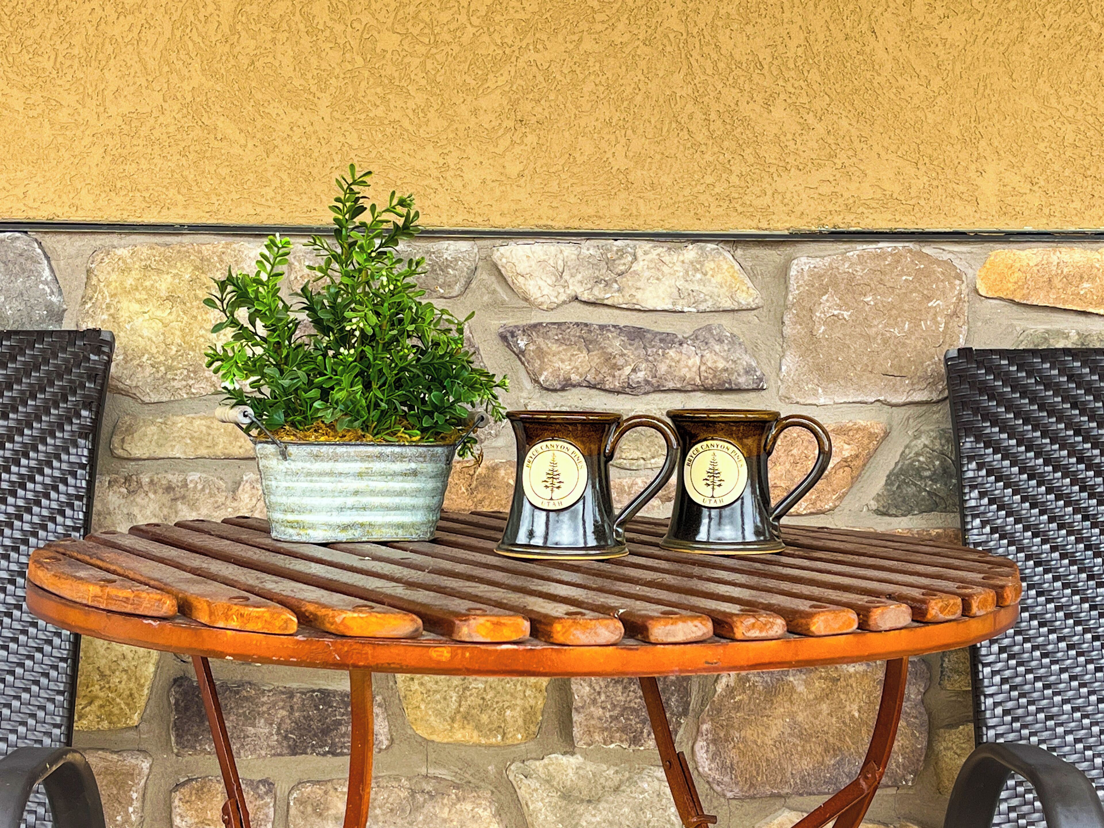 Una mesa de madera redonda con jarras de metal y una planta en maceta, frente a un muro de piedra.