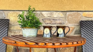 Una mesa de madera redonda con jarras de metal y una planta en maceta, frente a un muro de piedra.