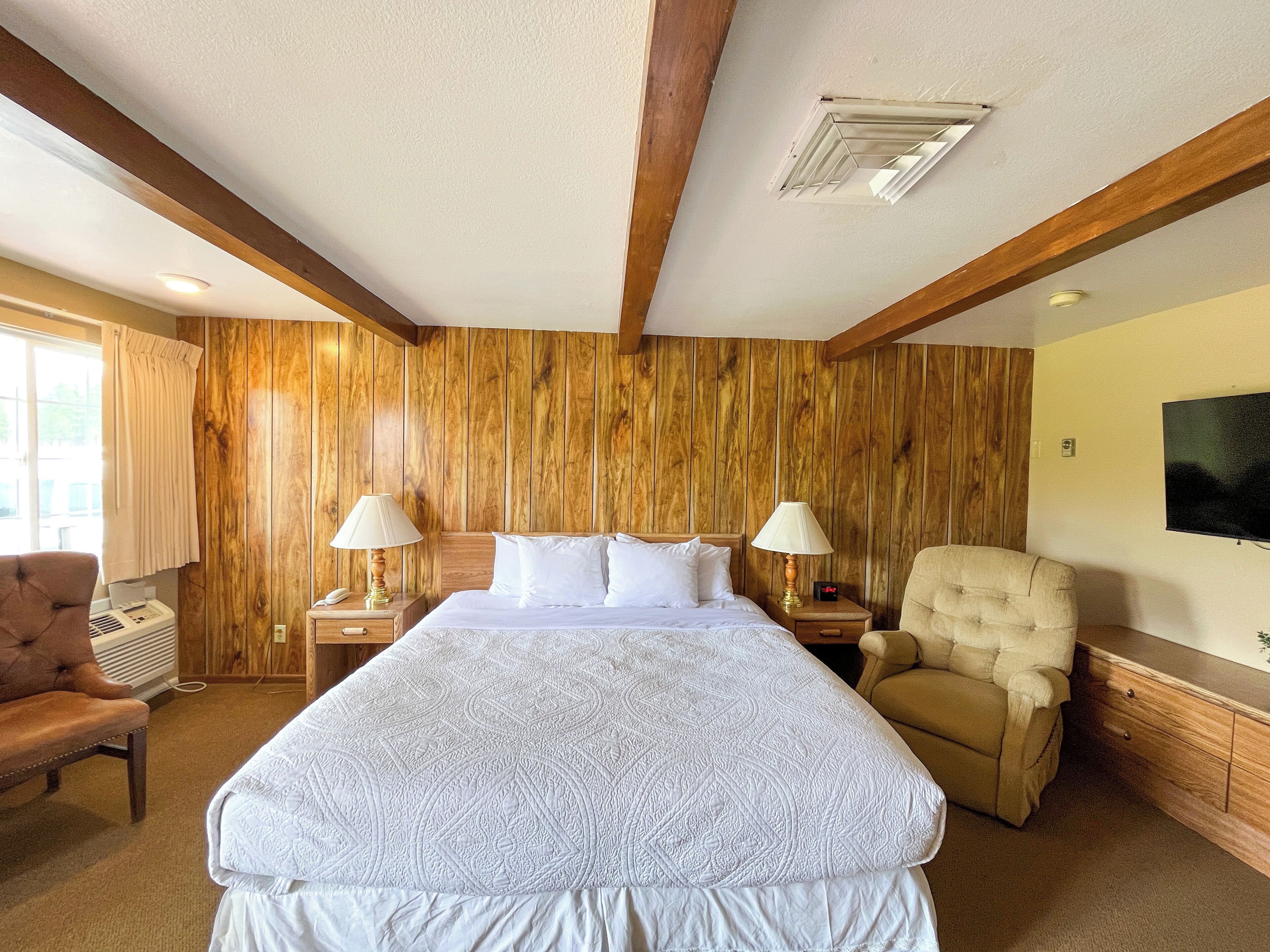 A hotel room with a large bed, two bedside tables with lamps, a wooden headboard, a beige armchair, and a television on the wall.