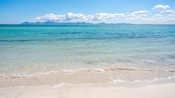 Een strand met helder water, een zandstrand en verre bergen onder een blauwe lucht.