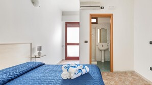 A bedroom with a bed, a bedside table with a lamp, a bathroom visible through an open door, and an air conditioning unit on the wall.