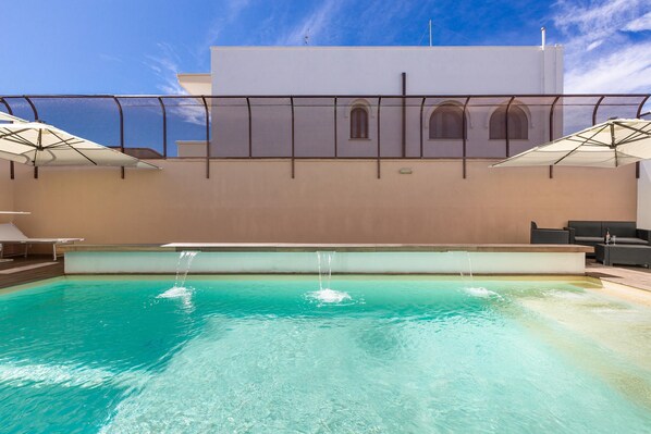 A swimming pool with clear blue water, a white building with arched windows, and outdoor seating with umbrellas.