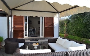 An outdoor patio with wicker furniture, a stone table with fruit, and a view of a garden.