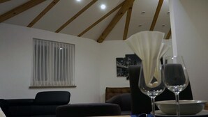 Una sala de estar con techo inclinado, una ventana con cortinas translúcidas, un sofá negro y una mesa de comedor con copas de vino y un cuenco.