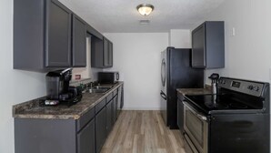 A compact kitchen with dark cabinets, a built-in microwave, a coffee maker, a stove, and a refrigerator. - Spacious West Side Home away from Home! (Wichita)