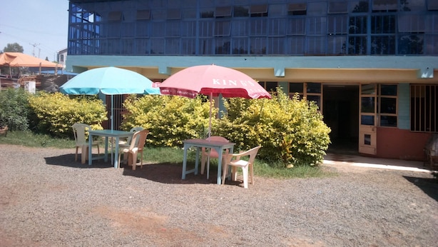 An outdoor seating area with two umbrellas, a gravel ground, and a building with a tiled roof and multiple windows. - Eden Garden Hotel (Wote)