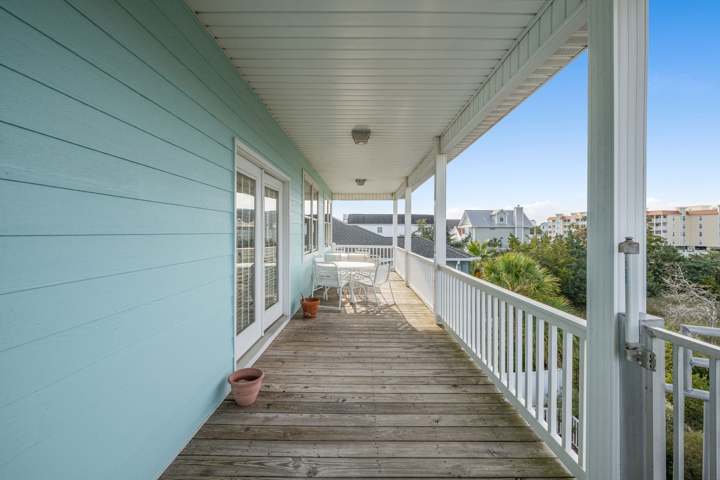 A wooden deck with white railings and a turquoise wall, leading to a covered area with a table and chairs.