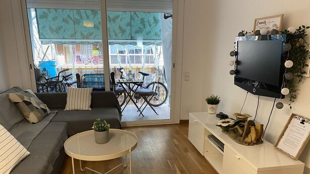 Un soggiorno con divano, un tavolino rotondo con una pianta, TV a muro e una porta vetrata scorrevole che dà su uno spazio esterno con biciclette.