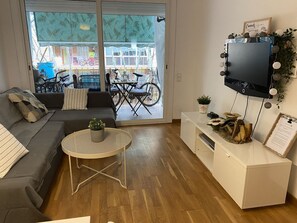 Ett vardagsrum med en soffa, ett litet runt bord med en växt, en tv som är monterad på väggen och en skjutdörr i glas som leder ut till ett utomhusområde med cyklar.