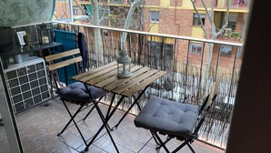 Un balcone con un tavolo di legno e due sedie, un piccolo ripiano con una figurina, e vista su un edificio con balconi e alberi.