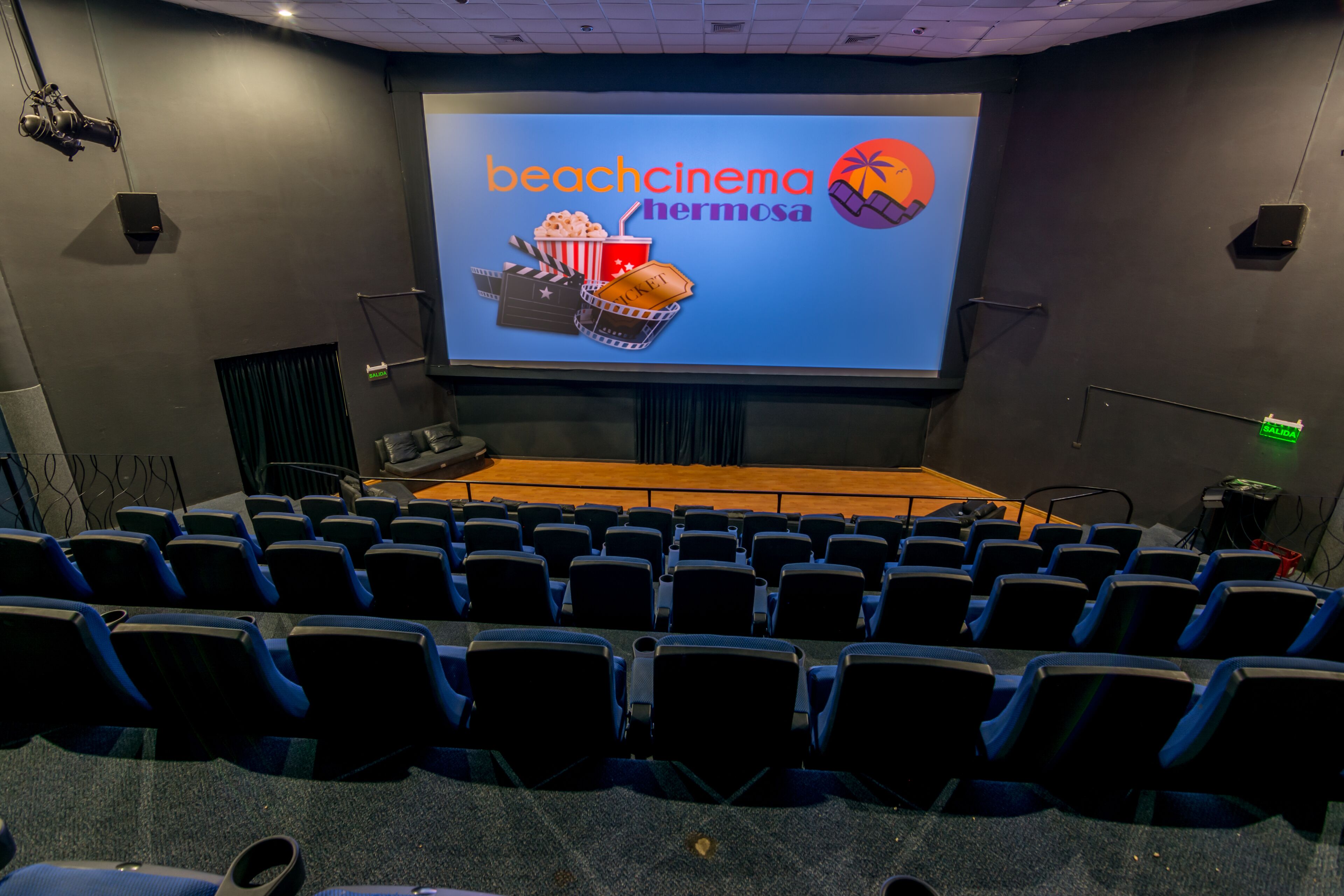 Un cinéma doté d’un grand écran sur lequel est affiché le texte « beachcinema hermosa », ainsi qu’un seau de pop-corn. Des rangées de sièges bleus sont disposées au premier plan.