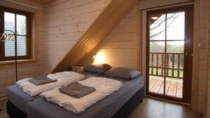 A bedroom with a sloped ceiling, a bed with pillows and blankets, a nightstand, a window, and a door leading to an outdoor deck. - Luxury Wooden Villa Albert in Forested Area with Stunning Countryside Views (Den Ham)