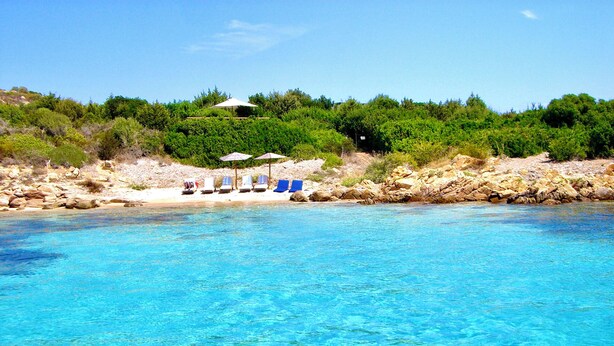 Une plage à l’eau d’un bleu limpide, bordée d’une rangée de transats et d’une côte rocheuse parsemée de végétation.