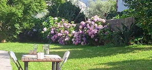 A garden with a table set for dining, surrounded by lush greenery and flowering bushes.