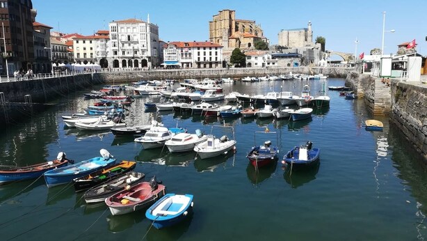 A marina with numerous boats docked, surrounded by historic buildings and a prominent stone structure in the background.