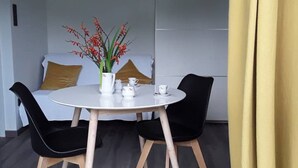 A modern room with a round table, two black chairs, a white sofa with a mustard-colored pillow, a vase with flowers, and a yellow curtain.