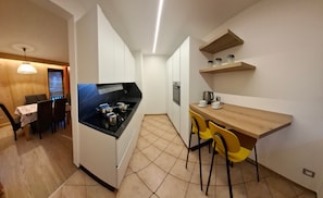 A compact kitchen with a black countertop, a built-in oven, and a coffee maker. There is a dining area with a table and chairs, and a wooden shelf above the counter. - Rustic Apartment Casa March 1 with Wi-Fi, Balcony, Garden & Mountain View (Moena)