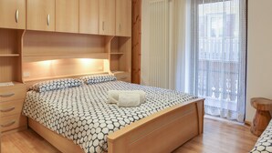 A bedroom with a wooden bed, a bedside table, a drawer unit, and a window with sheer curtains.