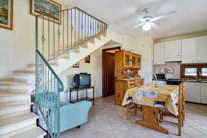 A kitchen with white cabinets, a dining table with a floral tablecloth, a wooden chair, a staircase with green railings, and a ceiling fan. - Holiday Home Villamara with Terrace, Mountain View & Garden; Parking Available (oliaspeciosa)
