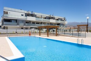 A swimming pool with a wooden pergola, a lifebuoy, and a clear view of a multi-story building with balconies in the background.